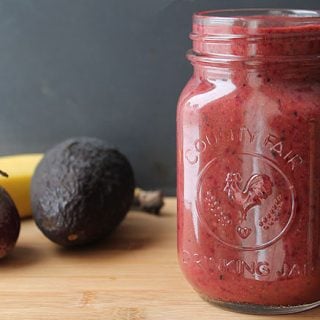 A red beet smoothie in a glass drinking jar, fresh avocados and bananas sit in the the background.