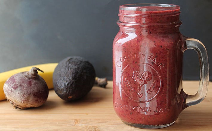 Berry Beet Ginger Smoothie in a glass on a wooden table with a fresh beet, avocado and banana sitting in the background.