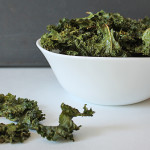 Easy Kale Chips in a white bowl, a few kale chips are in the forefront on the table.
