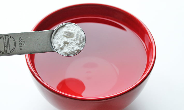 A measuring spoon holding baking powder over a bowl of water.