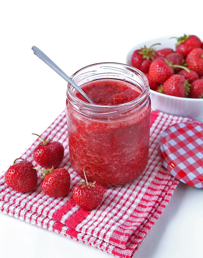 Strawberry Chia Seed Jam in a jar, jar is sitting on a white table and fresh strawberries are in front and behind the jar of jam.