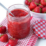 Strawberry Chia Seed Jam in a glass jar, a spoon is sticking out of the jar, the jar is sitting on a red and white dish cloth and there are fresh strawberries on and behind the cloth.