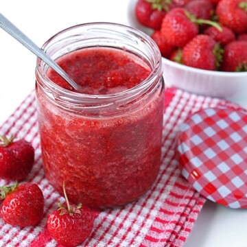Strawberry Chia Seed Jam in a glass jar, a spoon is in the jar, the jar is sitting on a red and white checkered dish cloth.