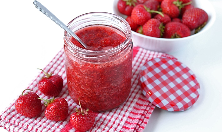 Strawberry Chia Seed Jam in a glass jar, a spoon is sticking out of the jar, the jar is sitting on a red and white dish cloth and there are fresh strawberries on and behind the cloth.