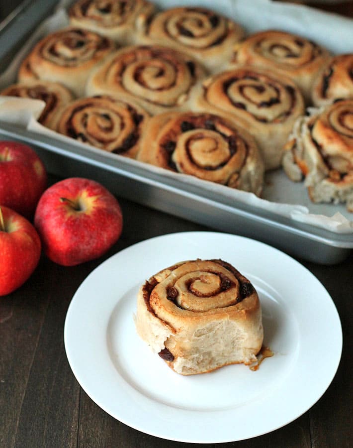 Vegan Apple Raisin Cinnamon Rolls in a pan in the background and one roll on a white plate in the foreground.