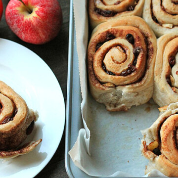 Vegan Apple Raisin Cinnamon Rolls sitting in a baking pan on the right and one roll is sitting on a white plate to the left.