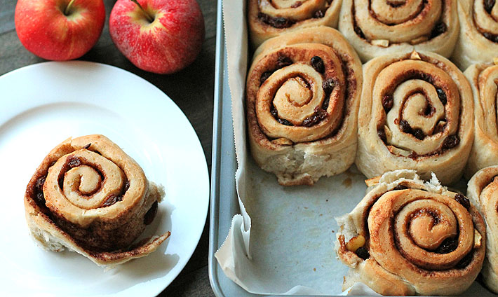 Vegan Apple Raisin Cinnamon Rolls sitting in a baking pan on the right and one roll is sitting on a white plate to the left.