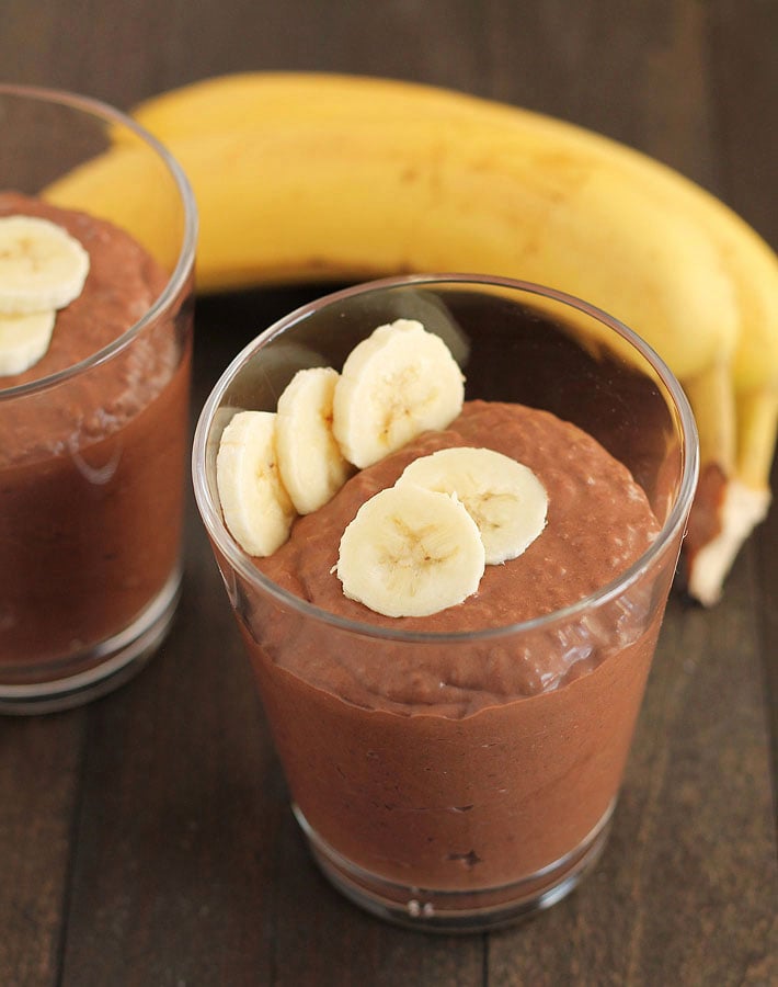 A clear cup filled with Chocolate Banana Coconut Chia Pudding sitting on a wooden table, two ripe bananas sit behind the cup.