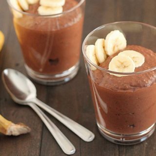Chocolate Banana Coconut Chia Pudding in a glass with sliced bananas on top for garnish, two spoons and two bananas sit to the left and another glass of pudding is behind.