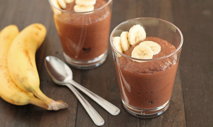 Chocolate Banana Coconut Chia Pudding in a glass with sliced bananas on top for garnish, two spoons and two bananas sit to the left and another glass of pudding is behind.