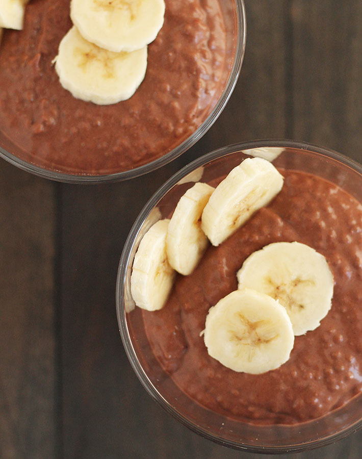 Overhead shot of two cups of Chocolate Banana Coconut Chia Pudding with sliced bananas on top, cups are sitting on a dark wooden table.