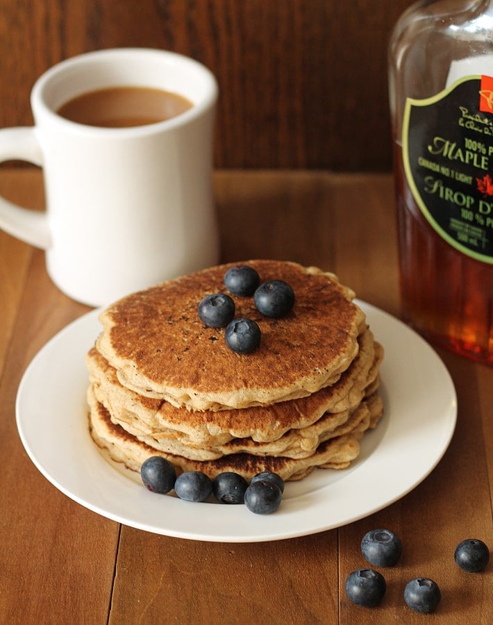 A stack of four Easy Vegan Gluten Free Pancakes on a white plate sitting on a brown wooden table, fresh blueberries are on top of the pancakes, a white mug of coffee sits in the background and a bottle of maple syrup sits to the right.