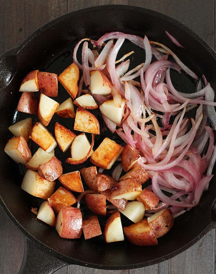 A cast iron pan with fried red potatoes and red onions that will be used to make a vegan breakfast burrito.