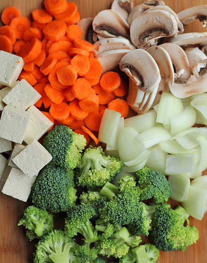 Chopped tofu, carrots, mushrooms, onions, and broccoli on a cutting board.