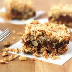 Three Vegan Date Squares on parchment squares on a brown wood table, a fork sits to the left.