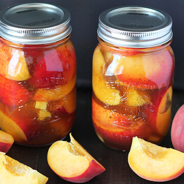 Two jars filled with pickled peaches, fresh peaches are on the table in front of the jars.