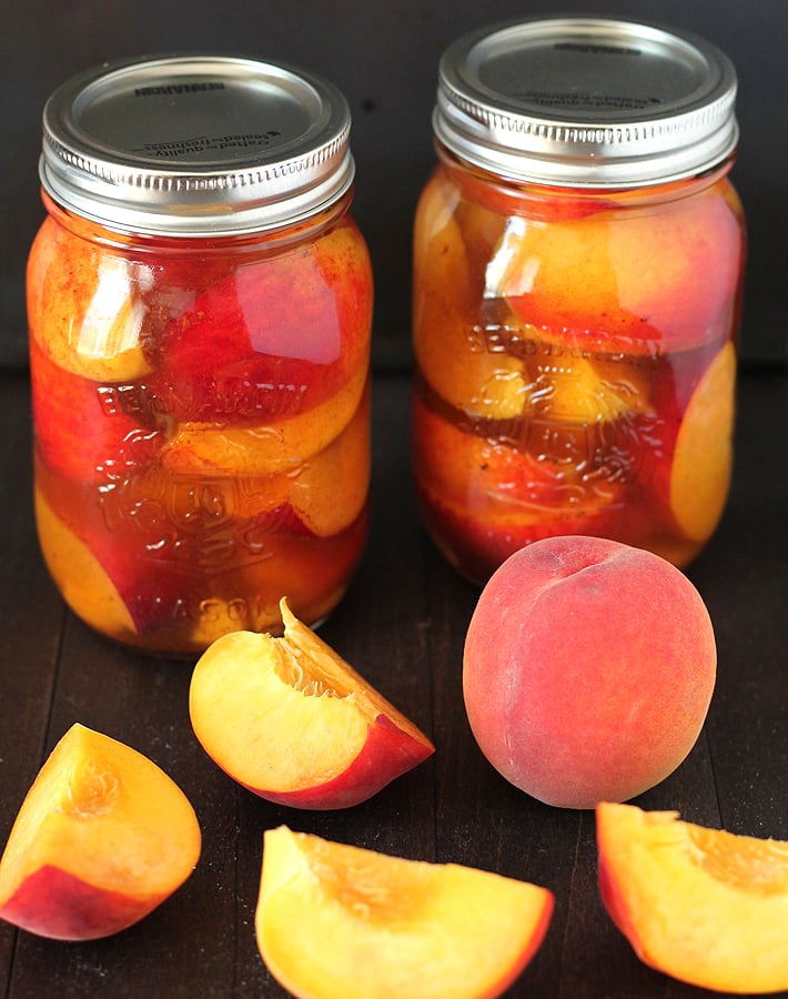 Two jars filled with pickled peaches, fresh peaches are on the table in front of the jars.