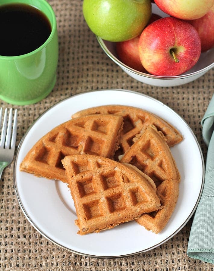 Picture of apple cinnamon waffles on a plate, a cup of coffee sits to the left and a bowl of apples sits behind the plate.