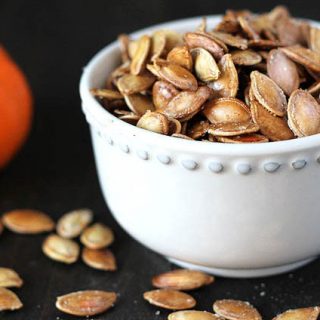 How to Roast Pumpkin Seeds - Roasted pumpkin seeds in a small white bowl on a dark wooden table, a small pumpkin is sitting off to the left of the bowl, some seeds are scattered on the table.