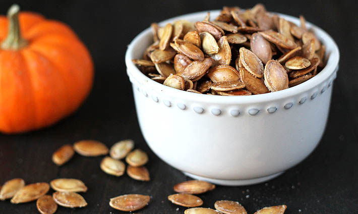 How to Roast Pumpkin Seeds - Roasted pumpkin seeds in a small white bowl on a dark wooden table, a small pumpkin is sitting off to the left of the bowl, some seeds are scattered on the table.