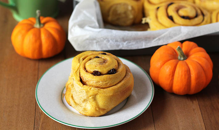 Vegan Pumpkin Cinnamon Rolls in a parchment lined baking pan with small pumpkins sitting on the side and in front of the pan.