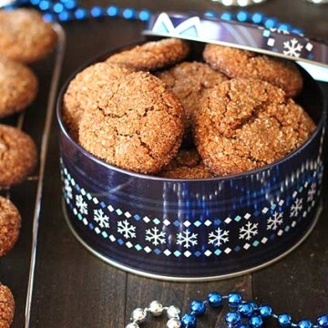 A tin of Vegan Gluten Free Gingerbread Cookies sitting beside silver and blue Christmas decorations and more cookies on a cooling rack.