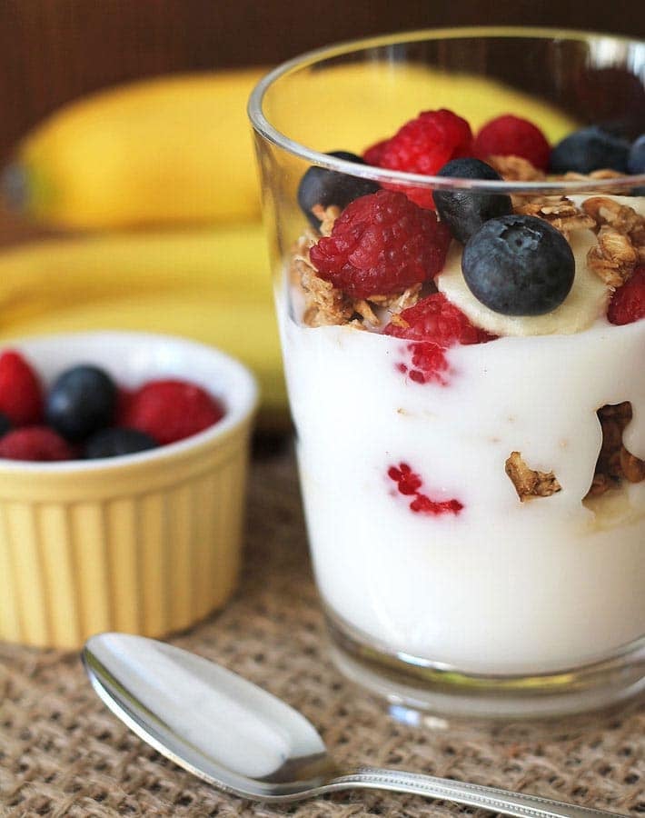 A vegan fruit and yogurt parfait on a placemat, a spoon sits in front of the parfait and a cup of berries sits to the left