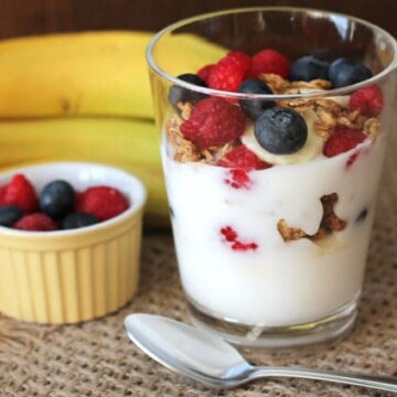 A fruit, yogurt, and granola parfait in a glass cup on a table with a spoon and fresh fruits.