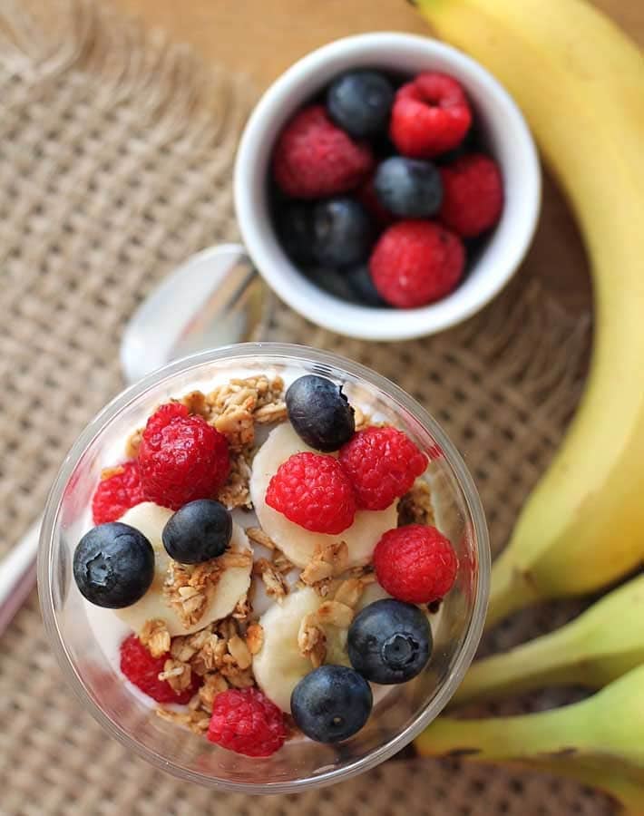 Overhead shot of a clear glass of Banana Berry Granola Yogurt Parfait a bunch of bananas and a cup of fresh berries, a spoon sits off to the left of the parfait.