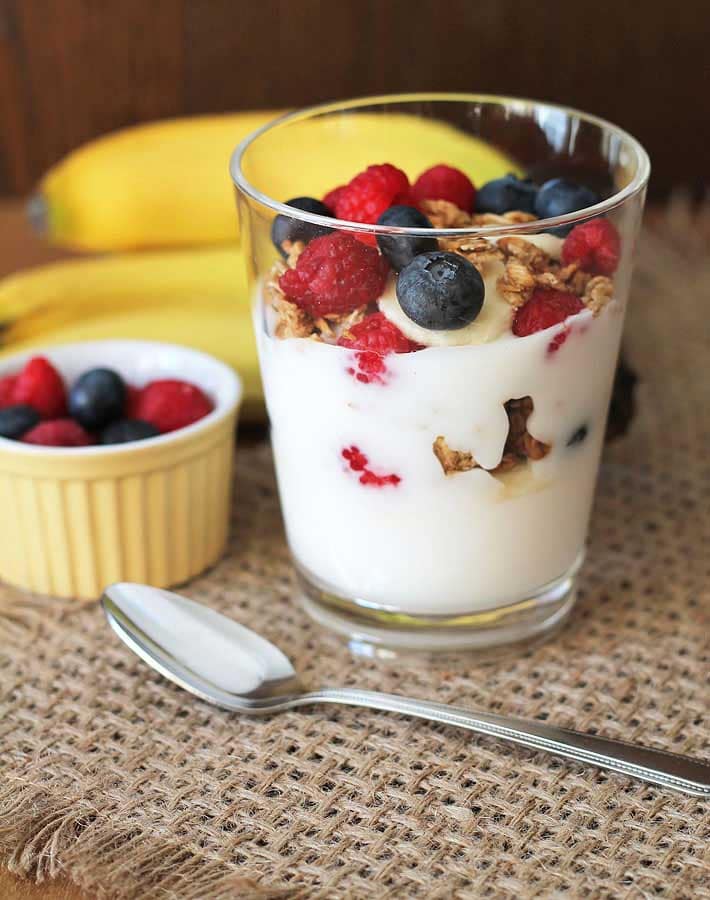 A Banana Berry Granola Yogurt Parfait in a clear glass sitting on a burlap placemat with a spoon in front of the glass, a bunch of bananas behind the glass and a small cup of berries on the left.
