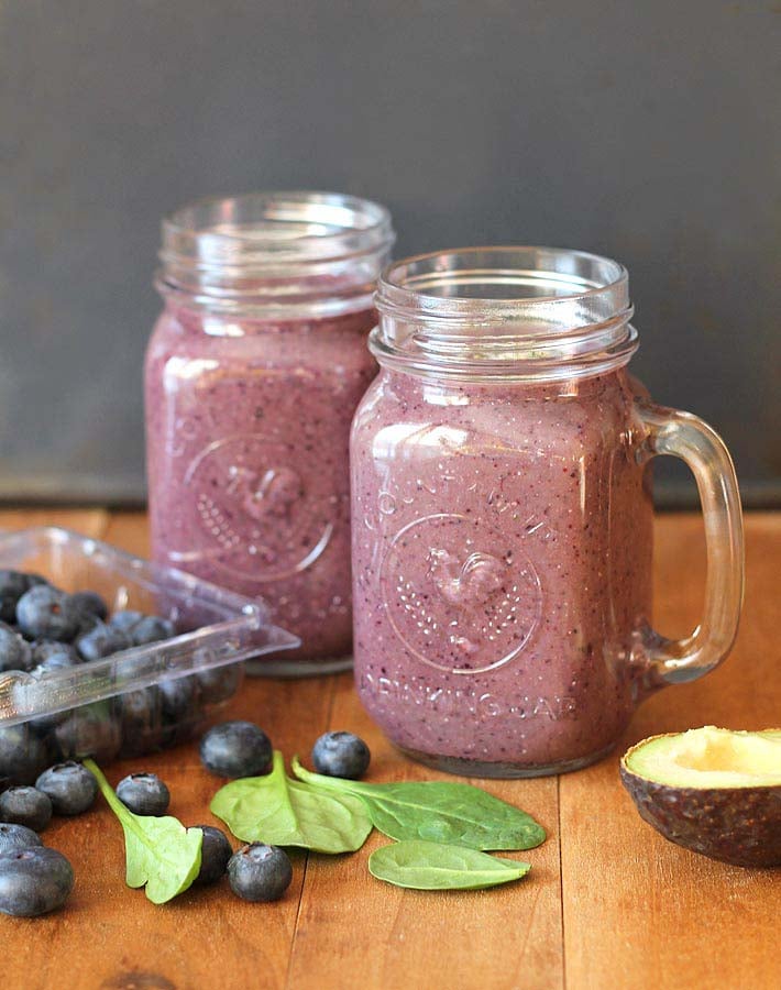 Blueberry Pineapple Smoothie in glass mugs on a table, fresh blueberries, avocando and spinach are also on the table.