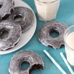 Vegan Gluten Free Baked Chocolate Doughnuts on a light blue table cloth, a white plate with three doughnuts sits to the back left, and a doughnut with a bite taken out of it is in the foreground with a glass of almond milk on the right.
