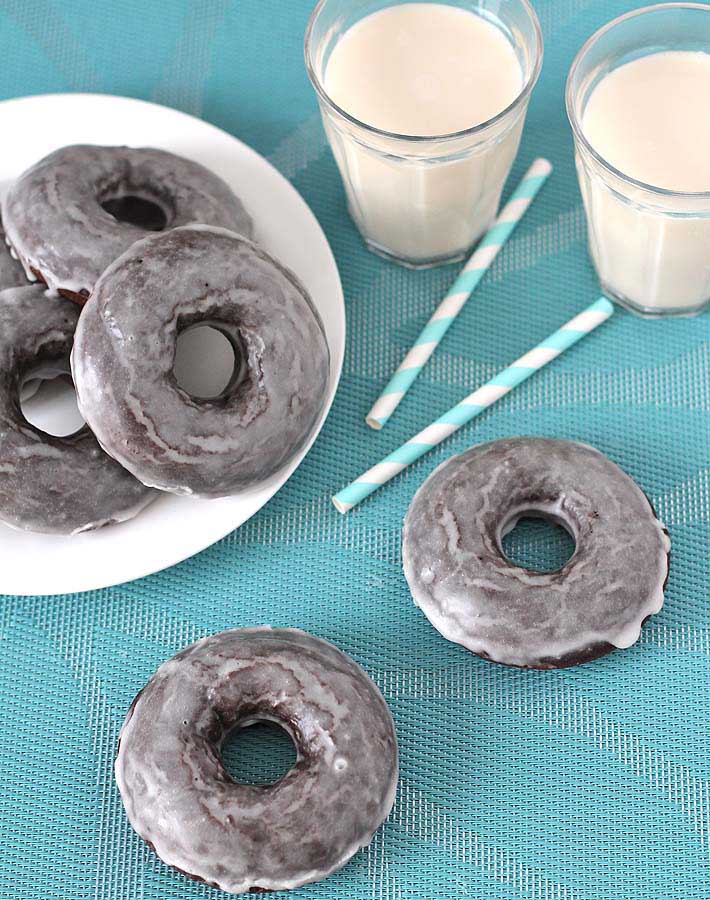 Vegan Gluten Free Baked Chocolate Doughnuts on a white plate on the left, two doughnuts are on a blue tablecloth and two glasses of almond milk sit behind the doughnuts with two paper straws in front of the glasses.