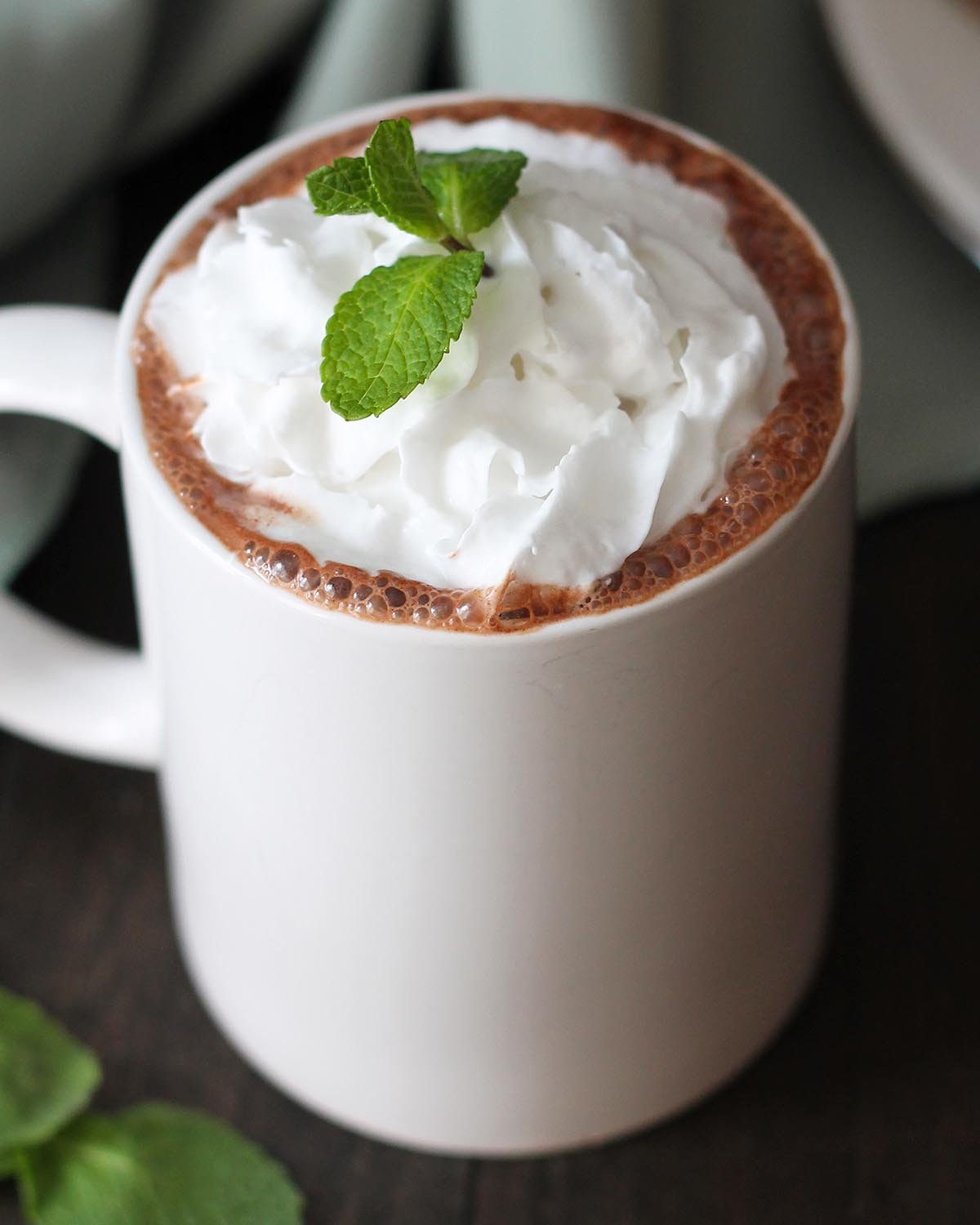 A mug of peppermint hot chocolate, it\'s garnished with whipped cream and a fresh mint leaf.