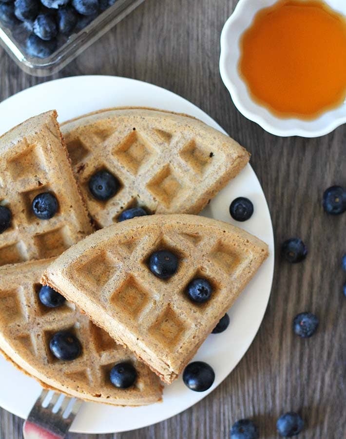 Overhead shot of Easy Vegan Gluten Free Waffles on a white plate sitting on a wood table, waffles are garnished with fresh blueberries, a container of blueberries sits behind the plate and a cup of maple syrup sits to the right.