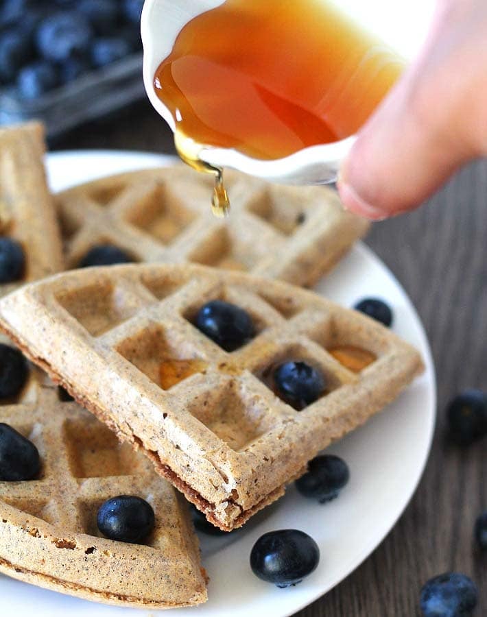 Maple syrup in a small white ceramic cup being poured onto a plate of Easy Vegan Gluten Free Waffles, waffles are garnished with fresh blueberries.