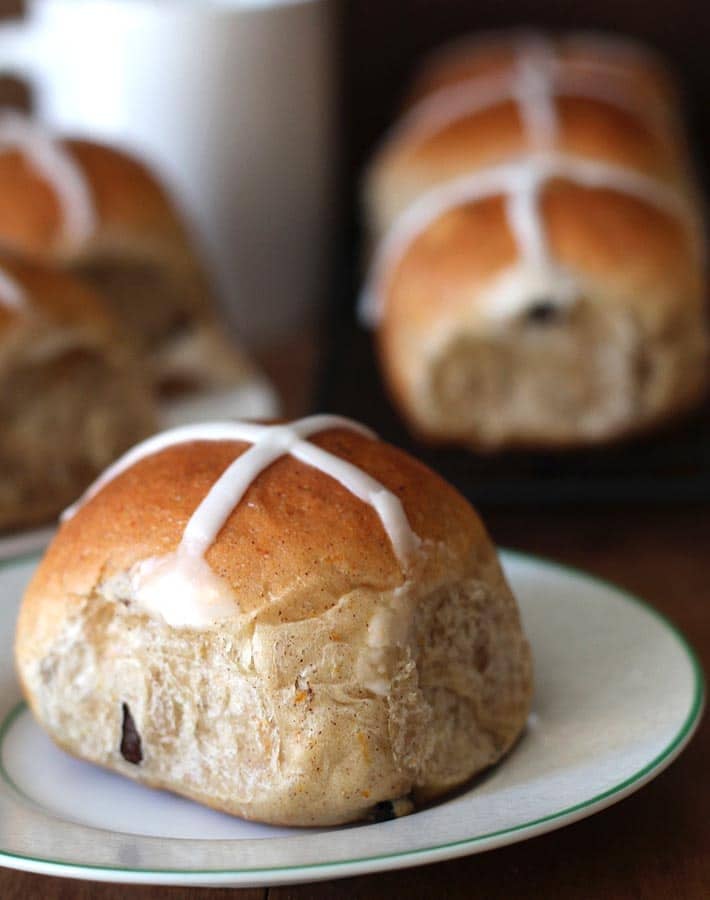 An up close shot of Vegan Hot Cross Buns, one is sitting on a plate while the others are sitting behind it.