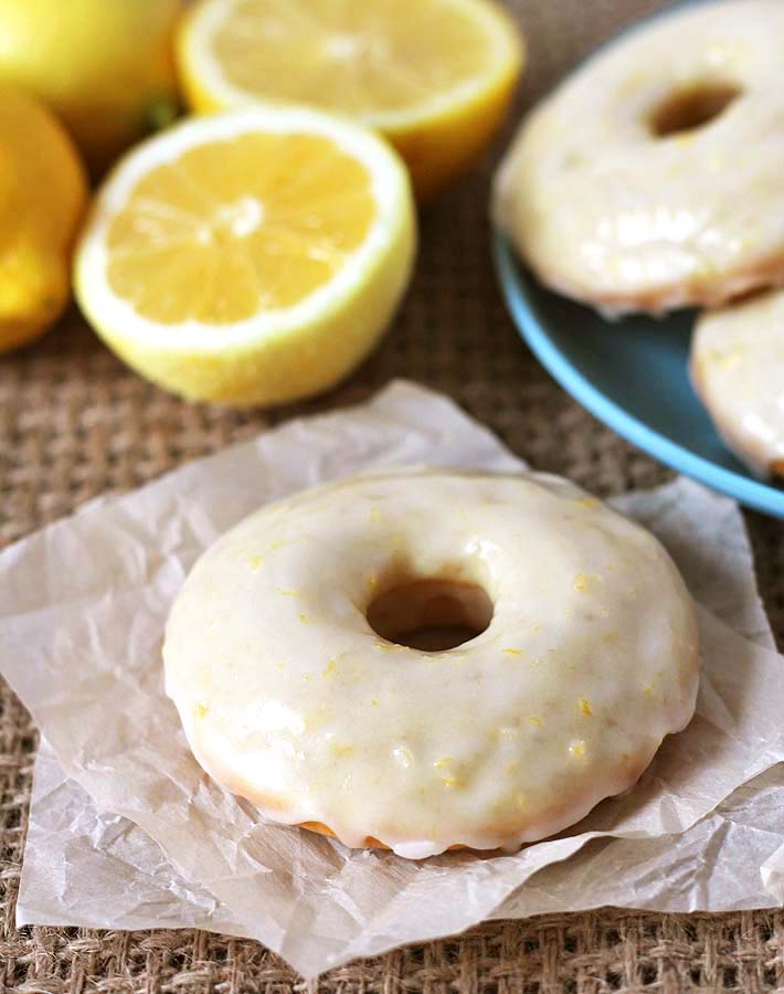 Vegan Baked Lemon Doughnuts on a blue plate to the right inthe background, one doughnut sits on parchment paper in the foregound, fresh sliced lemons sit in the background.