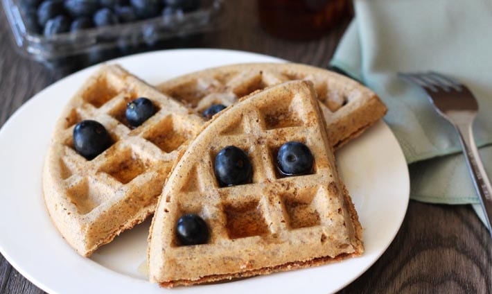 Easy Vegan Gluten Free Waffles on a white plate, a green cloth napkin sits to the right with a fork on top of it, waffles are garnished with fresh blueberries, and a container of blueberries sits behind the plate.