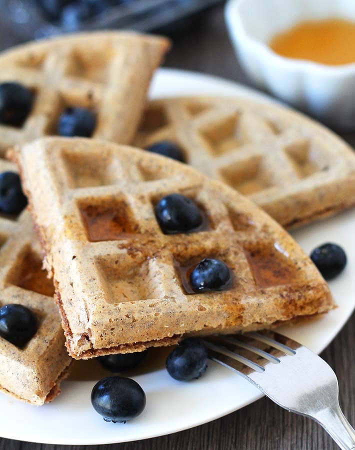 Vegan gluten free waffles on a white plate with fresh blueberries and syrup on top, a fork sits on the side of the plate.