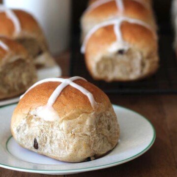 Vegan Hot Cross Buns sitting on a light brown wooden table, one bun sitting on a plate in the forefront.