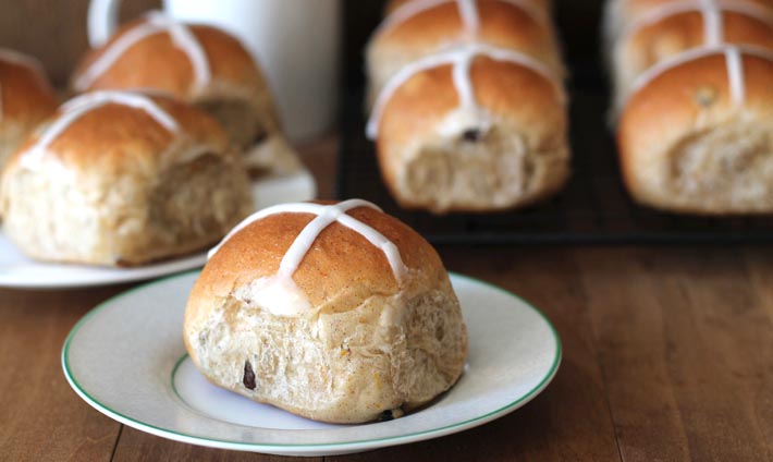 Vegan Hot Cross Buns sitting on a light brown wooden table, one bun sitting on a plate in the forefront.