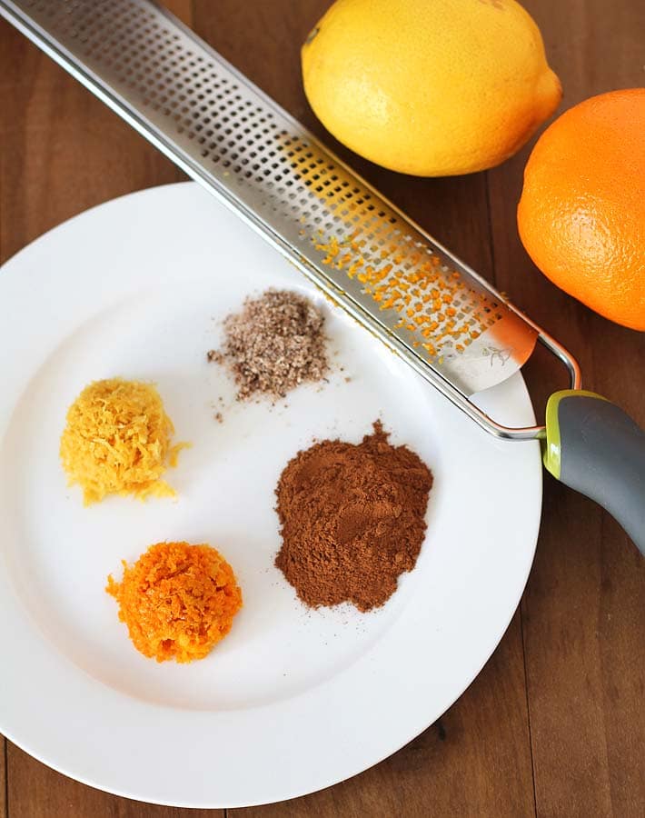 The spices and flavourings for Vegan Hot Cross Buns on a white plate (orange zest, lemon zest, nutmeg, and cinnamon).