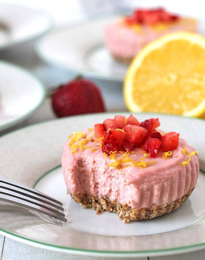 A Vegan Lemon Strawberry Cheesecake Bites sitting a plate with a piece of it missing and other bites sitting on plates in the background
