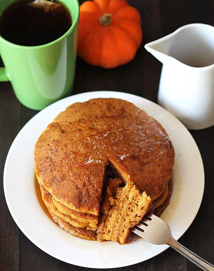 A stack of pumpkin pancakes sitting on a white plate with a cup of tea to the left and a cup of syrup on the right 