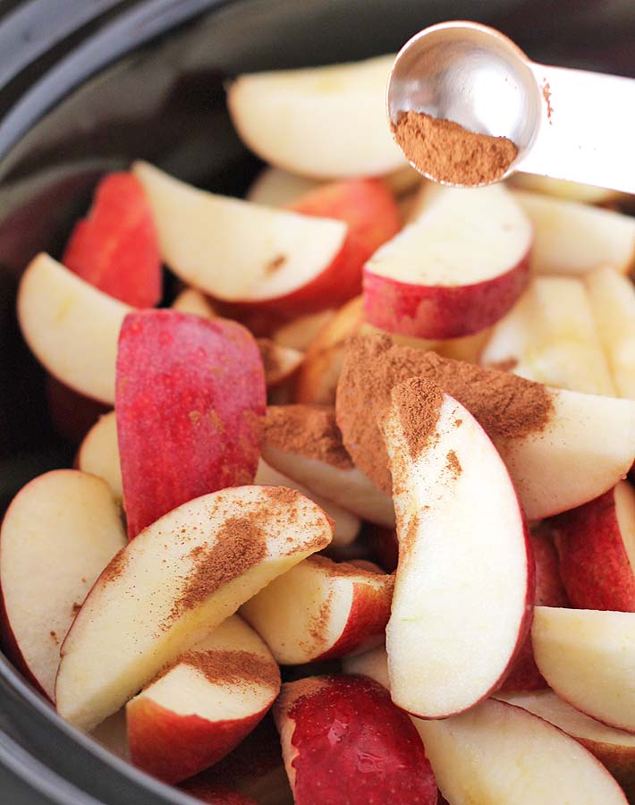 Freshly cut apples in a slow cooker with cinnamon being sprinkled on them to prepare them to make slow cooker applesauce