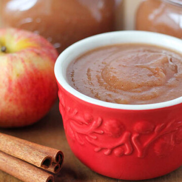 a cup of slow cooker applesauce with cinnamon sticks, a whole apple, and two jars of applesauce on a table