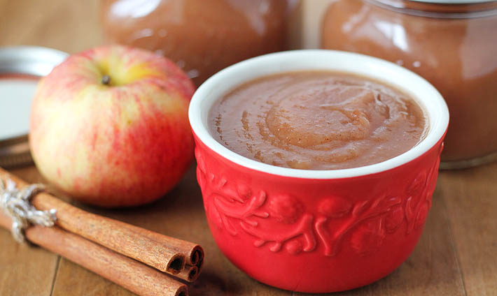 a cup of slow cooker applesauce with cinnamon sticks, a whole apple, and two jars of applesauce on a table