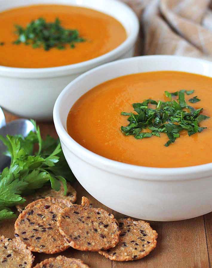 Two bowls of Butternut Squash Sweet Potato Carrot Soup sitting on a brown wooden table with crackers in the front of one bowl and fresh parsley on the left.