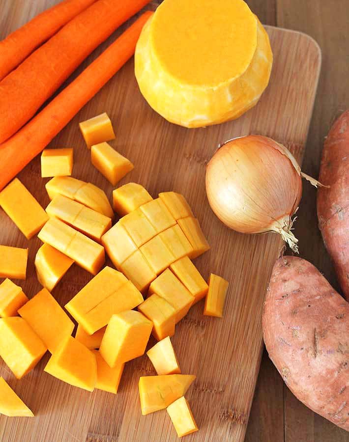 A cutting board with chopped butternut squash, three peeled carrots, the bottom of a butternut squash, and two unpeeled sweet potatoes on the right to make Butternut Squash Sweet Potato Carrot Soup.